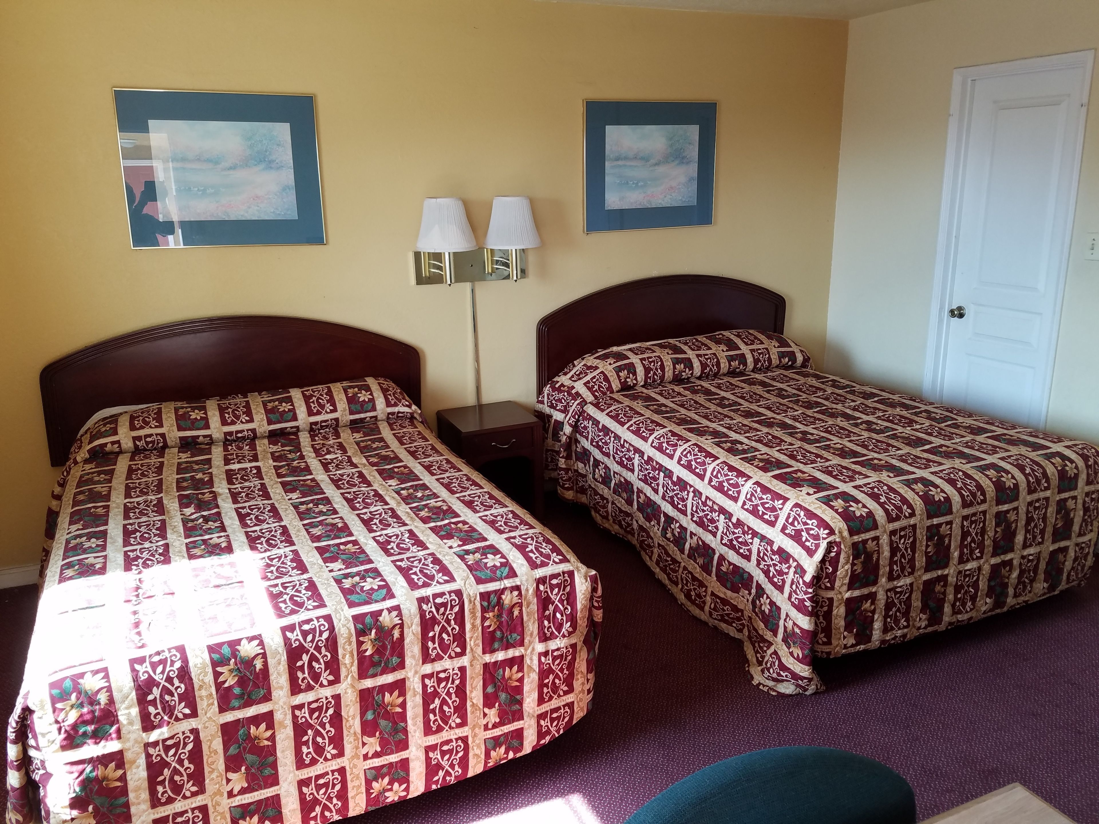 Habitación de hotel con dos camas, cada una con una colcha estampada, una mesita de noche con lámpara y dos cuadros enmarcados en la pared.