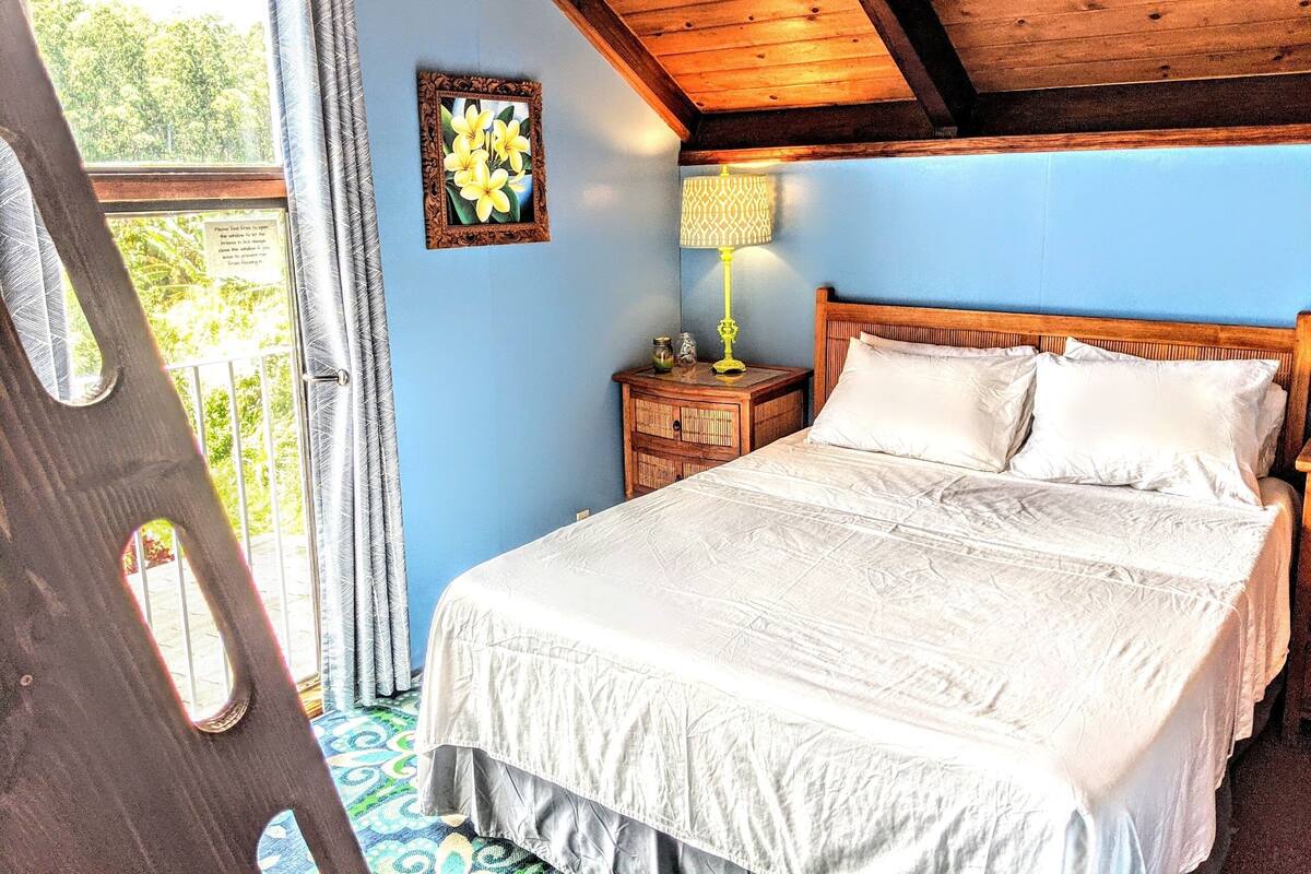 Ein Schlafzimmer mit Holzboden, einem Bett mit weißer Bettwäsche, einem Nachttisch mit Lampe und einem Fenster mit Vorhängen.