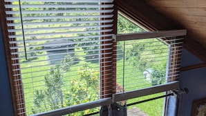 Une fenêtre avec des stores blancs, donnant sur un jardin où l’on peut voir différentes plantes, et une structure en arrière-plan.