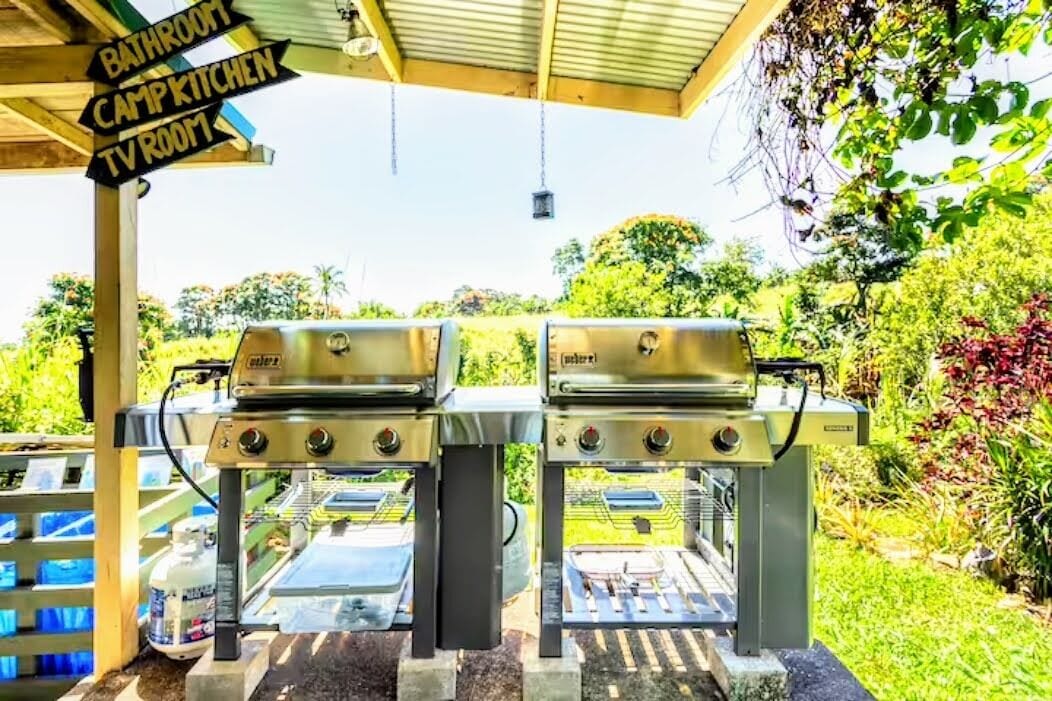 Duas churrasqueiras ao ar livre sob uma área coberta, com placas indicativas para 'Banheiro', 'Cozinha de Camping' e 'Sala de TV'.