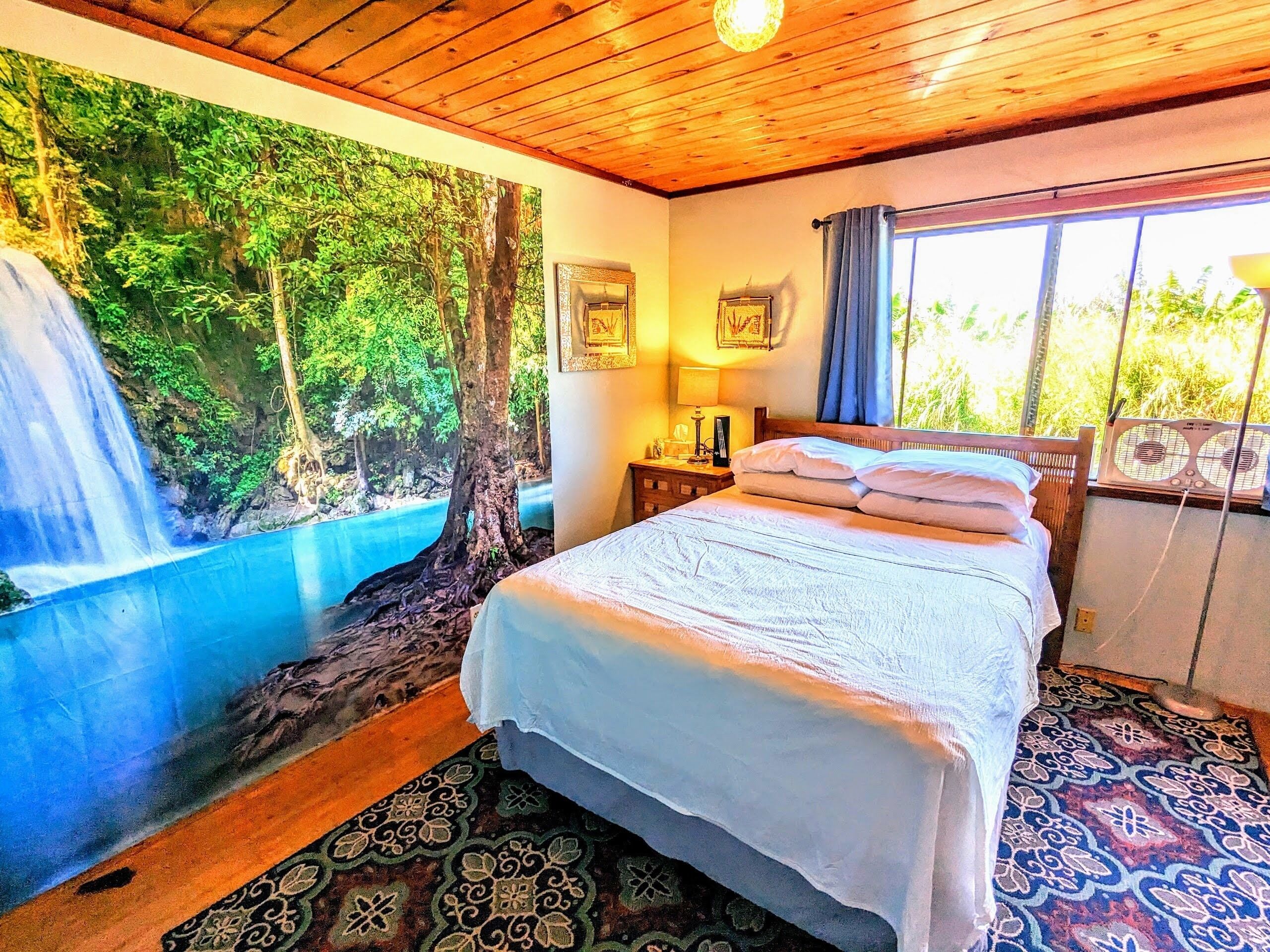 A bedroom with a bed, a nightstand, a wall-mounted lamp, a window with curtains, and a mural of a forest scene.