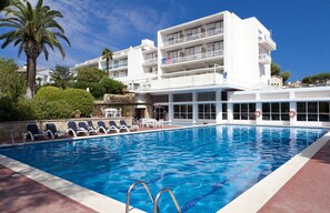 A swimming pool with lounge chairs, a palm tree, and a multi-story building with balconies. - Hotel Alga (Palafrugell)
