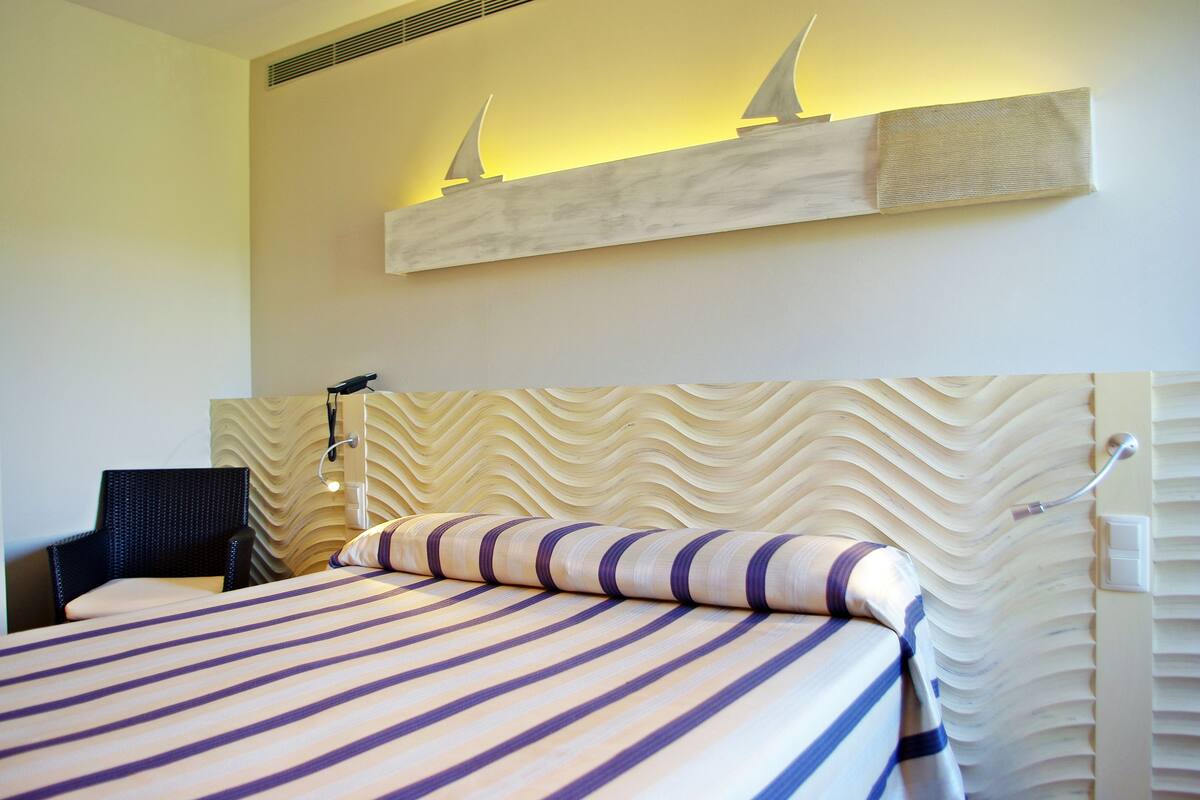 A hotel room with a bed, a chair, and a wall-mounted shelf with two sailboat decorations.