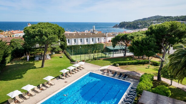 Piscina con sillas de descanso y sombrillas, rodeada de vegetación y con vistas a la costa.