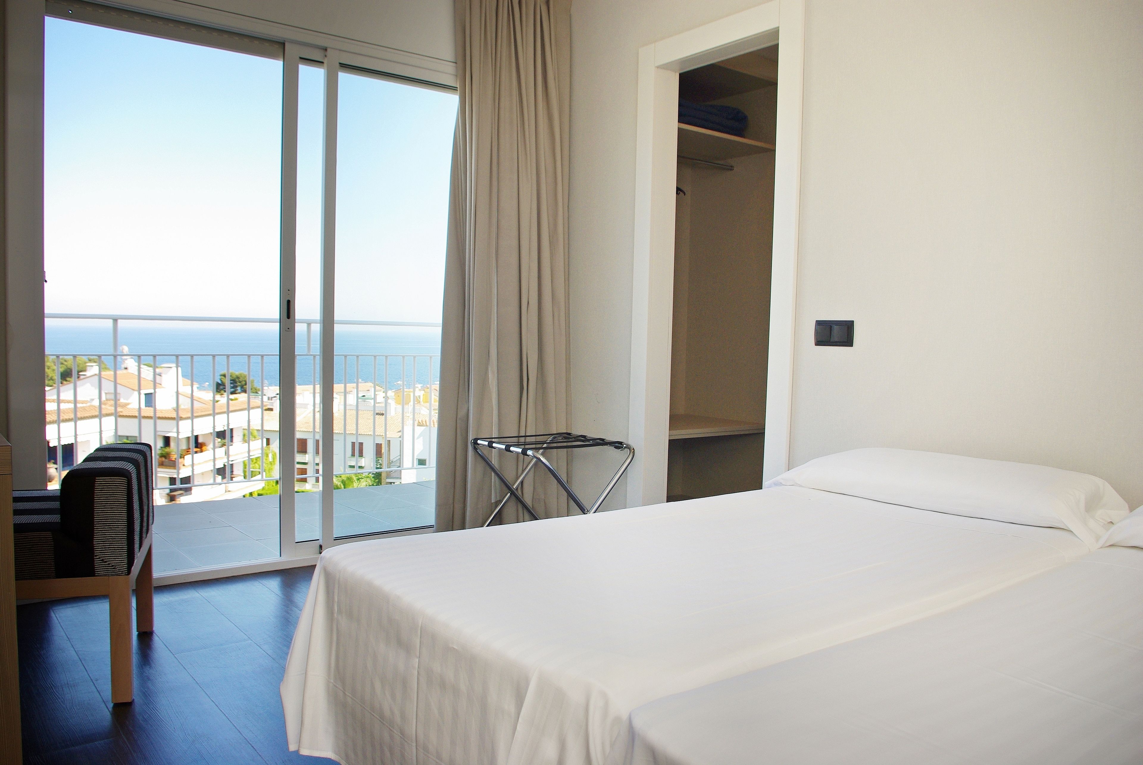 A hotel room with a bed, a seating area with a chair, a drying rack, and a view of the sea and buildings.