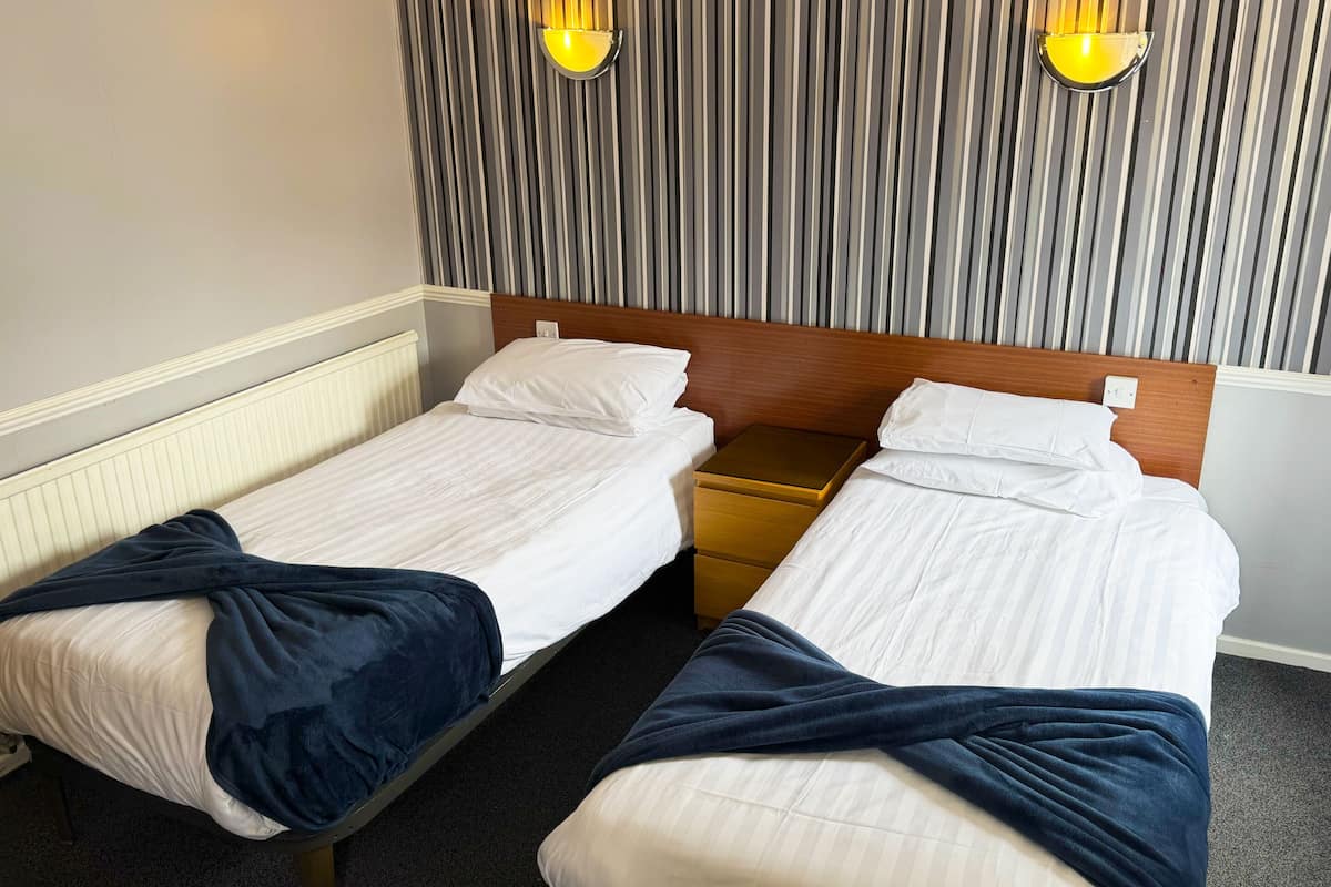 A hotel room with two single beds, each with a wooden headboard and white bedding. There are two bedside tables, one on each bed, and a striped wall panel above the beds.