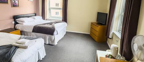 A hotel room with two beds, a TV, a desk, a chair, a fan, and a window with curtains.