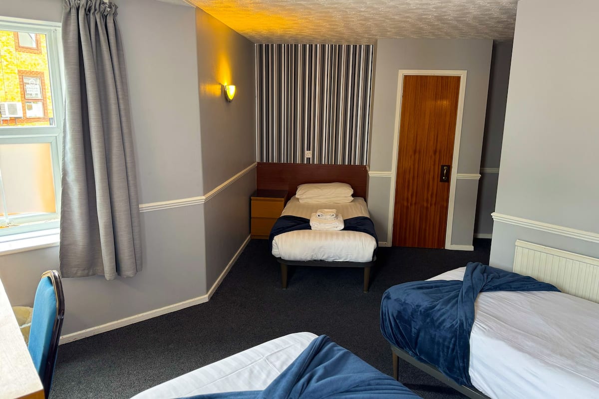 A hotel room with two single beds, a small bedside table, a window with curtains, a striped wall panel, and a door.