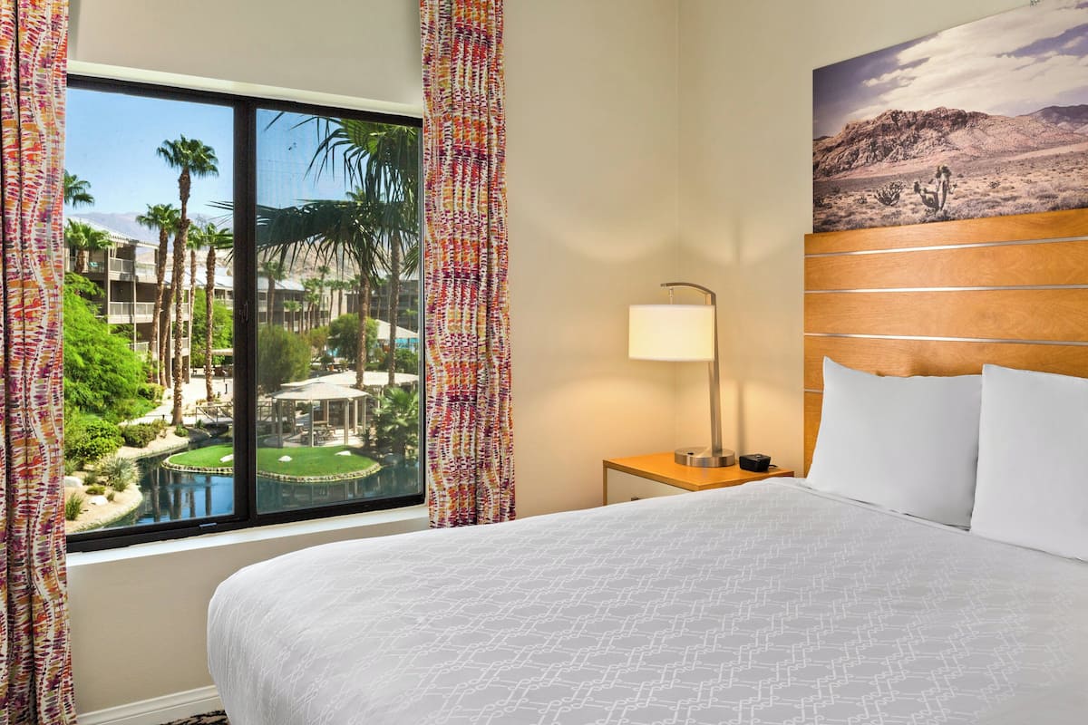 A hotel room with a large bed, a nightstand with a lamp, a view of a landscaped garden through a window, and a painting on the wall.