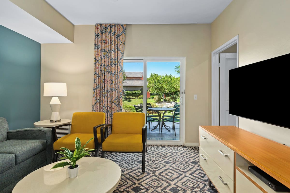 A hotel room with a sofa, a yellow armchair, a TV, a lamp, and a view of the outdoors through a glass door.