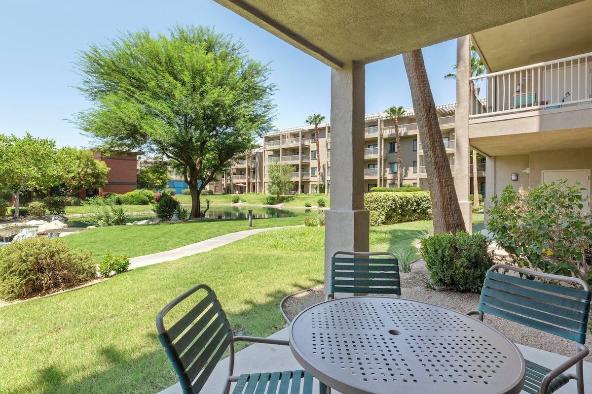 A courtyard with a round table and chairs, surrounded by greenery and a building with balconies.