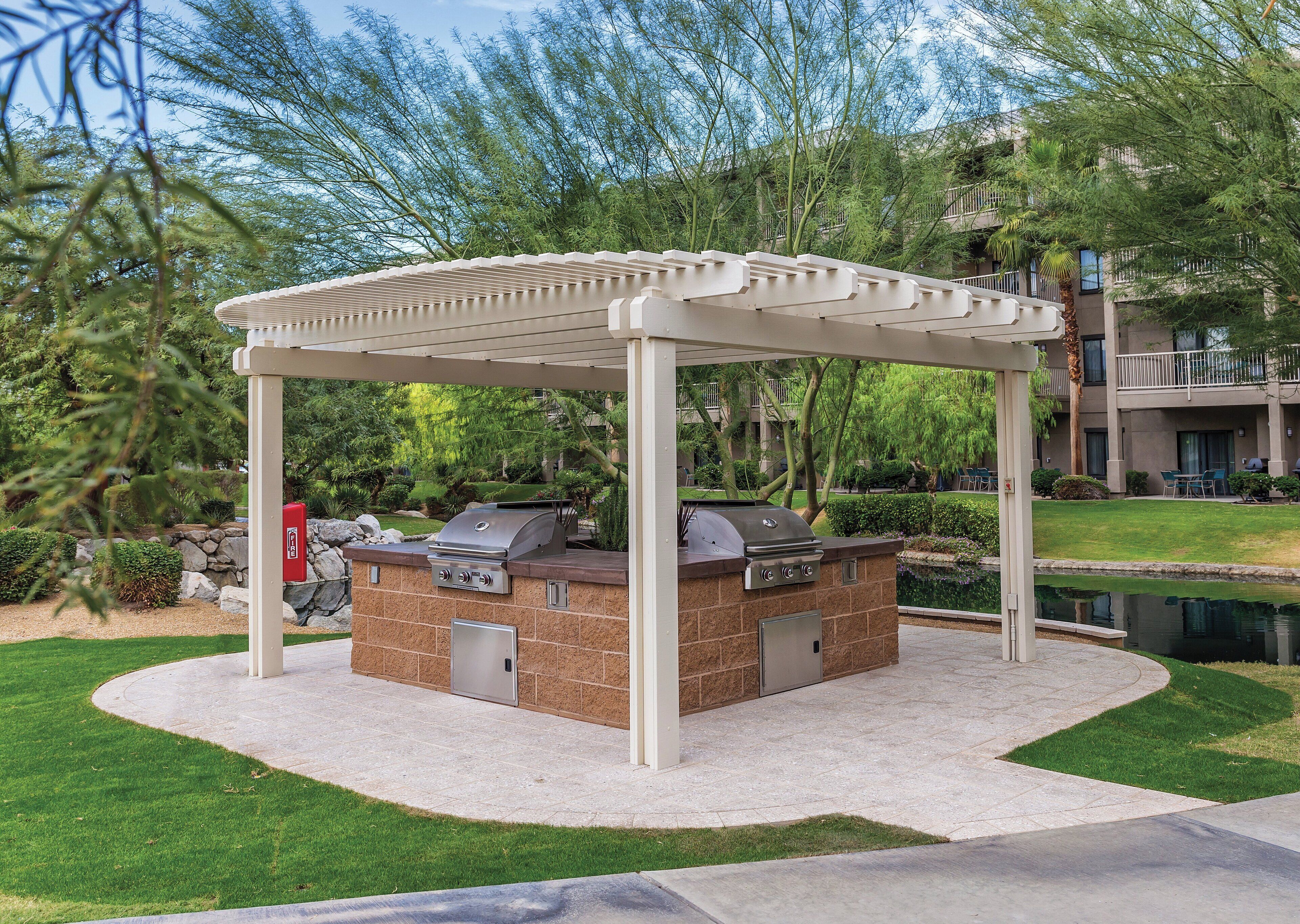 An outdoor barbecue area with a grill and a covered seating area, surrounded by greenery and a building in the background.
