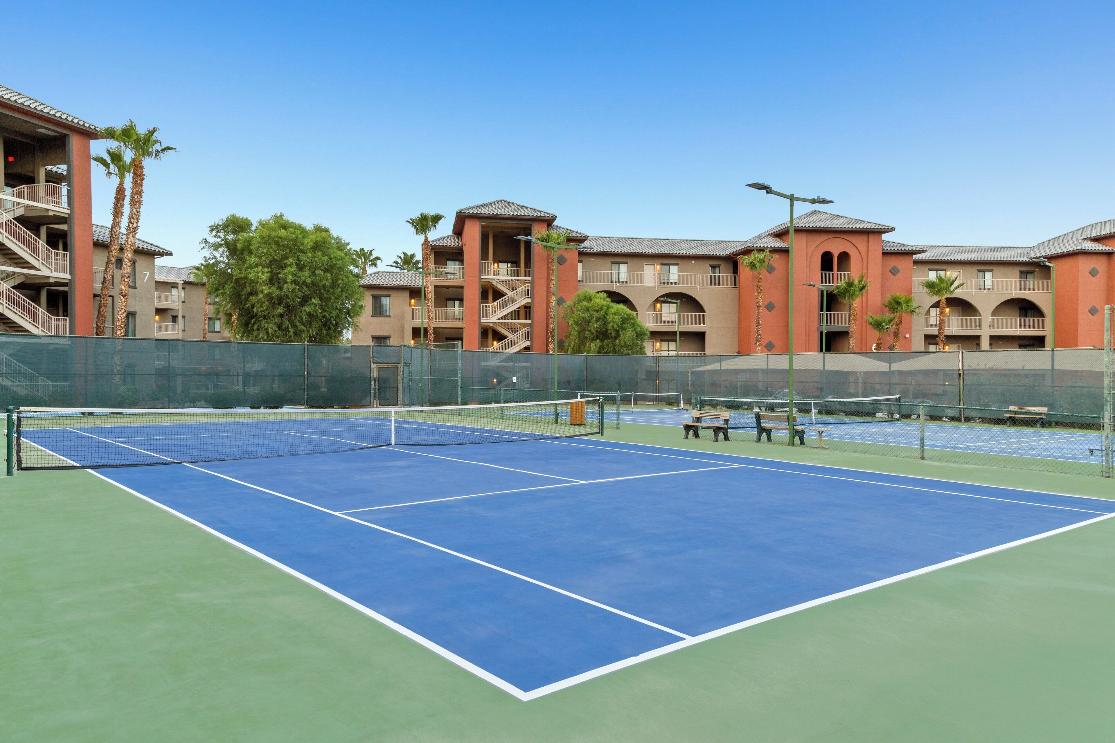 A tennis court with a net, surrounded by a fence, and adjacent to a building with multiple balconies and palm trees.