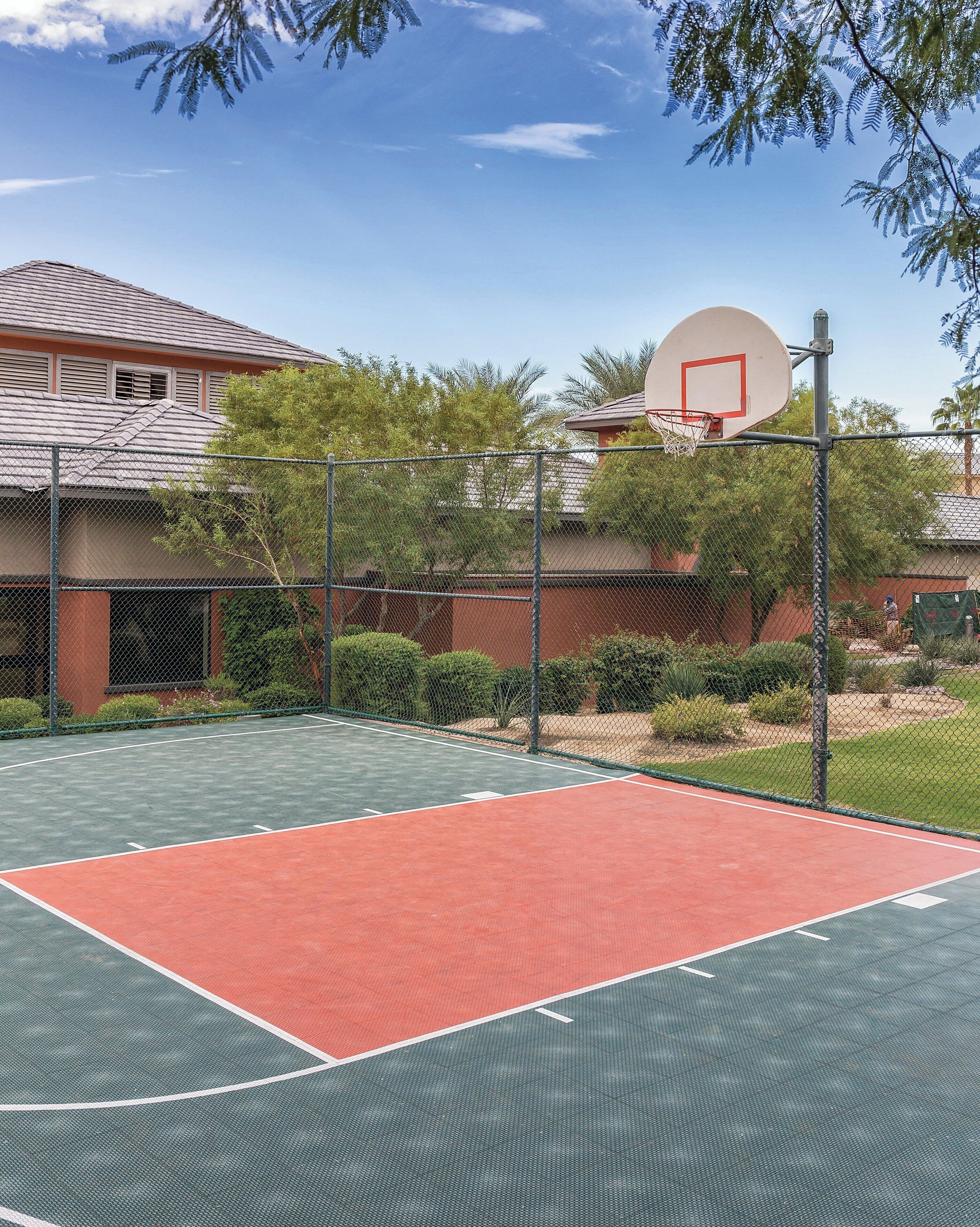 An outdoor basketball court with a chain-link fence, surrounded by trees and a building in the background.