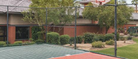 An outdoor basketball court with a chain-link fence, surrounded by trees and a building in the background.