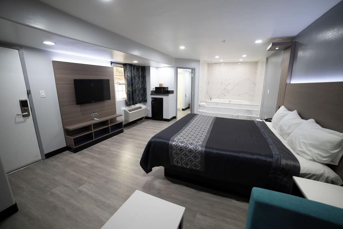 A modern hotel room with a large bed, a flat-screen TV, a built-in air conditioning unit, a small seating area, and a bathtub.