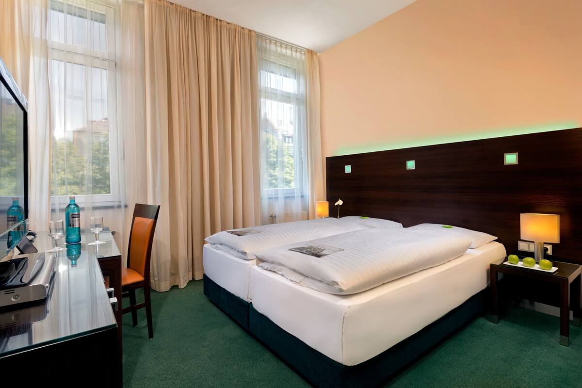 Habitación de hotel con una cama grande, un escritorio con teléfono y una botella de agua, una silla y una mesita de noche con lámpara.