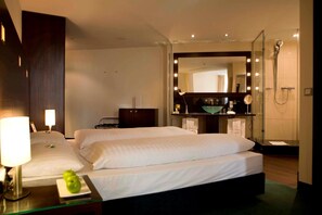 A hotel room with two beds, a bedside table with a lamp, a green apple, a mirror, a sink, and a shower. - Flemings Hamburger Allee (Frankfurt)