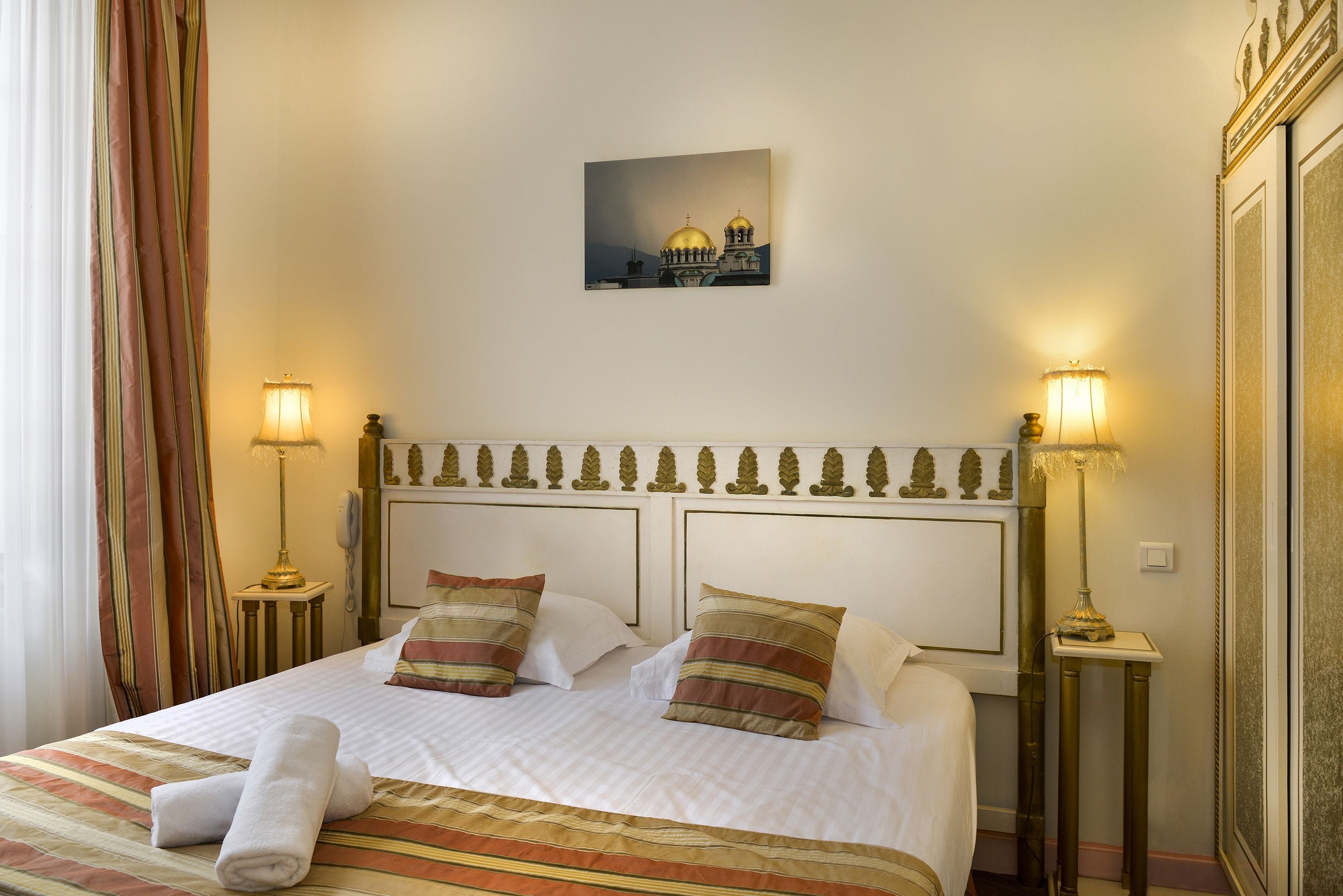 Una camera d'albergo con un letto rifatto, due abat-jour, un quadro incorniciato al muro e una finestra con tende.