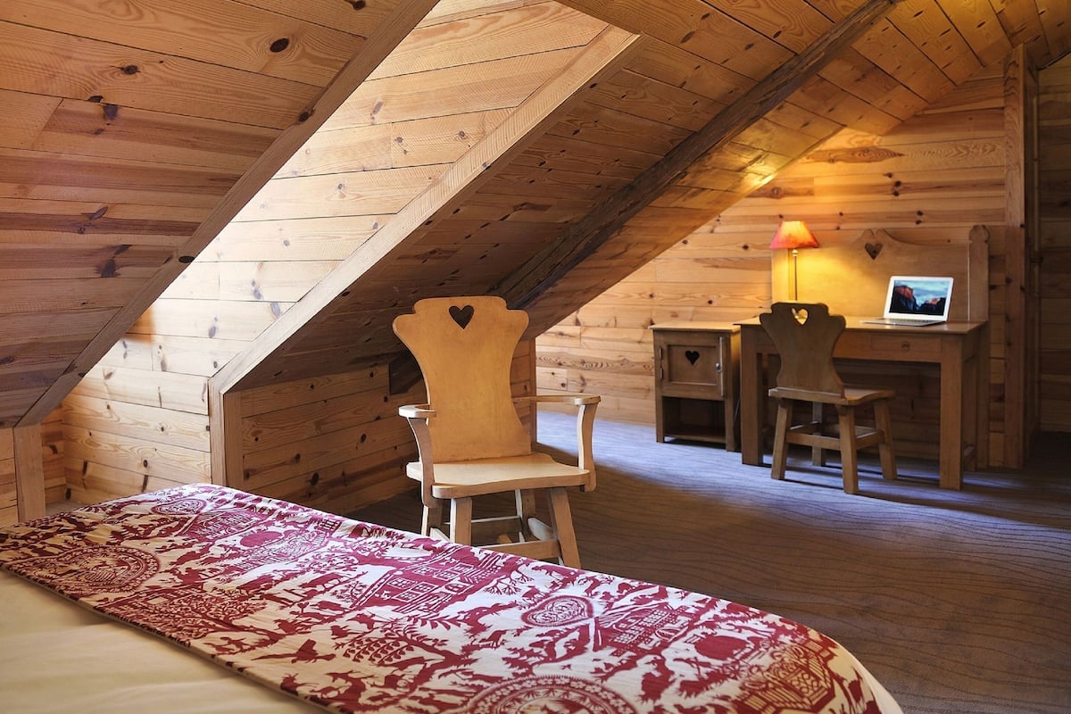 Een knus slaapkamer op zolder met een schuine kap, een bed, twee stoelen, een bureau met een laptop en een lamp.