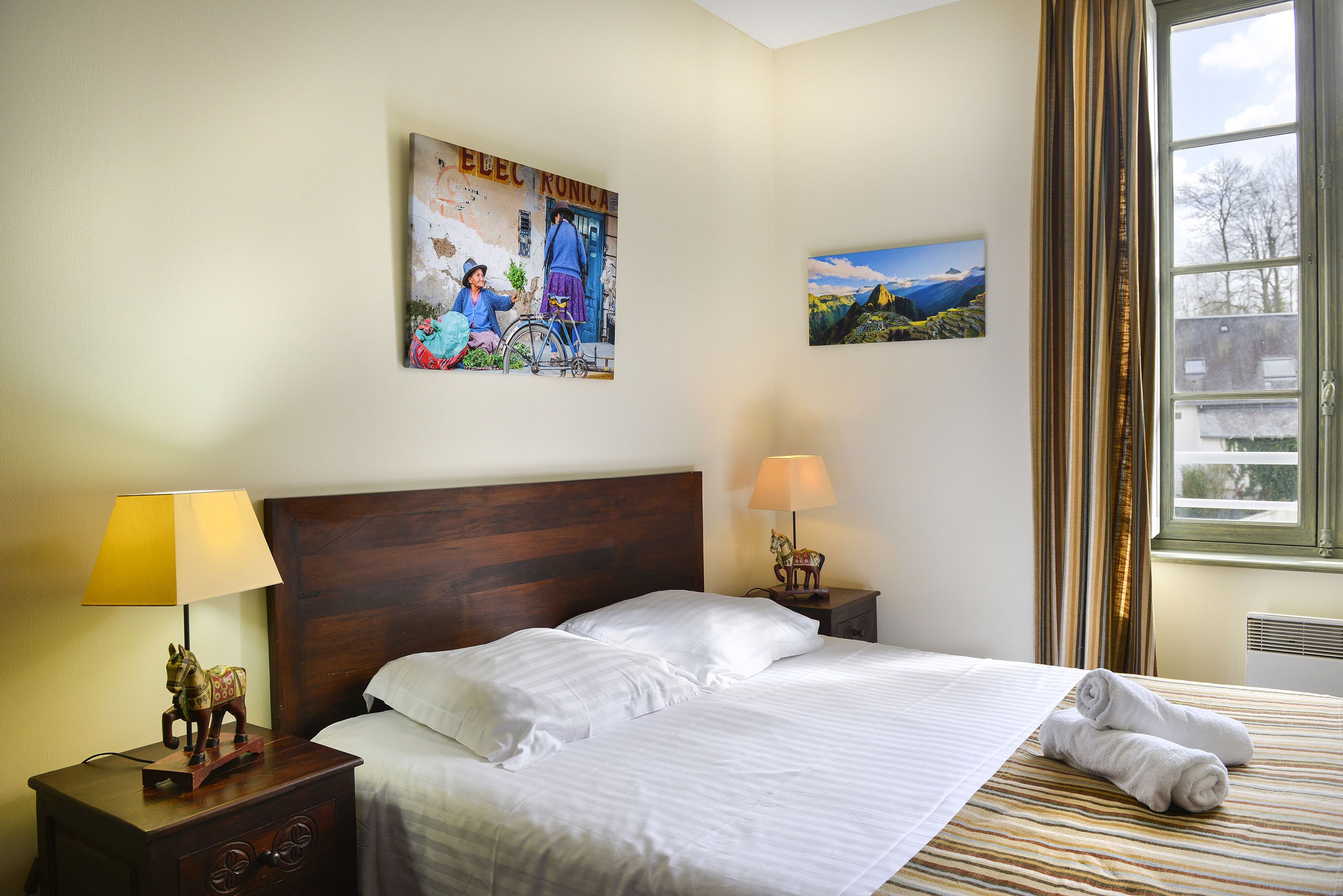 A neatly made bed with white linens, a wooden headboard, and a lamp on a bedside table. There are two paintings on the wall, one depicting a rural scene and the other a mountainous landscape.