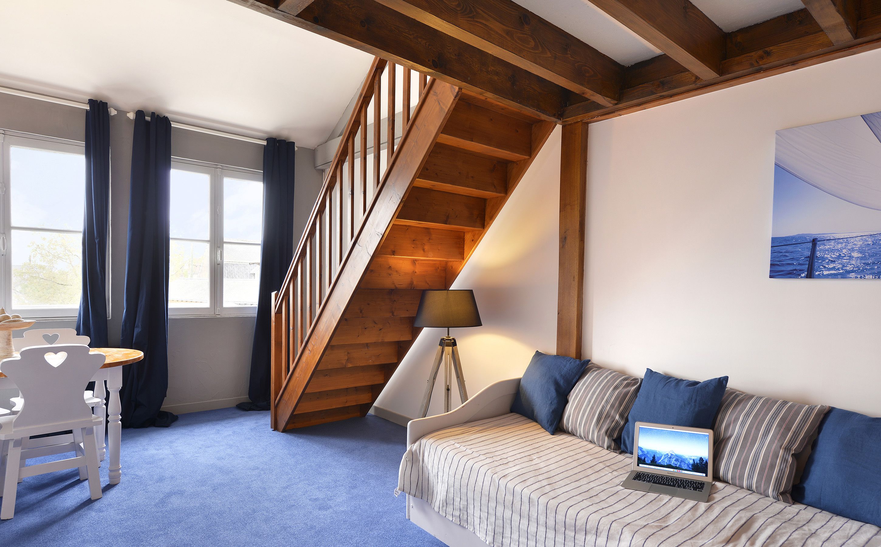 Un dormitorio con escalera de madera, cama con una laptop, escritorio y ventana con cortinas.