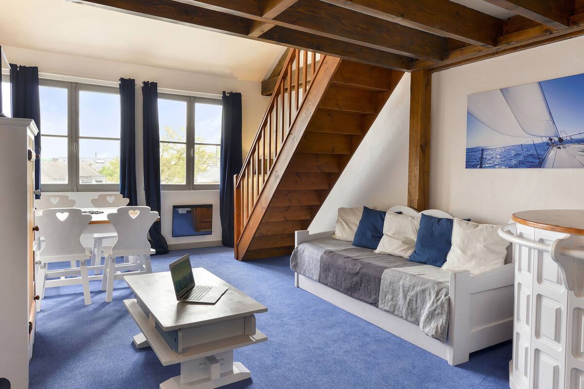 Een gezellige kamer met een houten trap, een bed met blauw-witte beddengoed, een bureau met een laptop en uitzicht op het omliggende landschap door grote ramen.