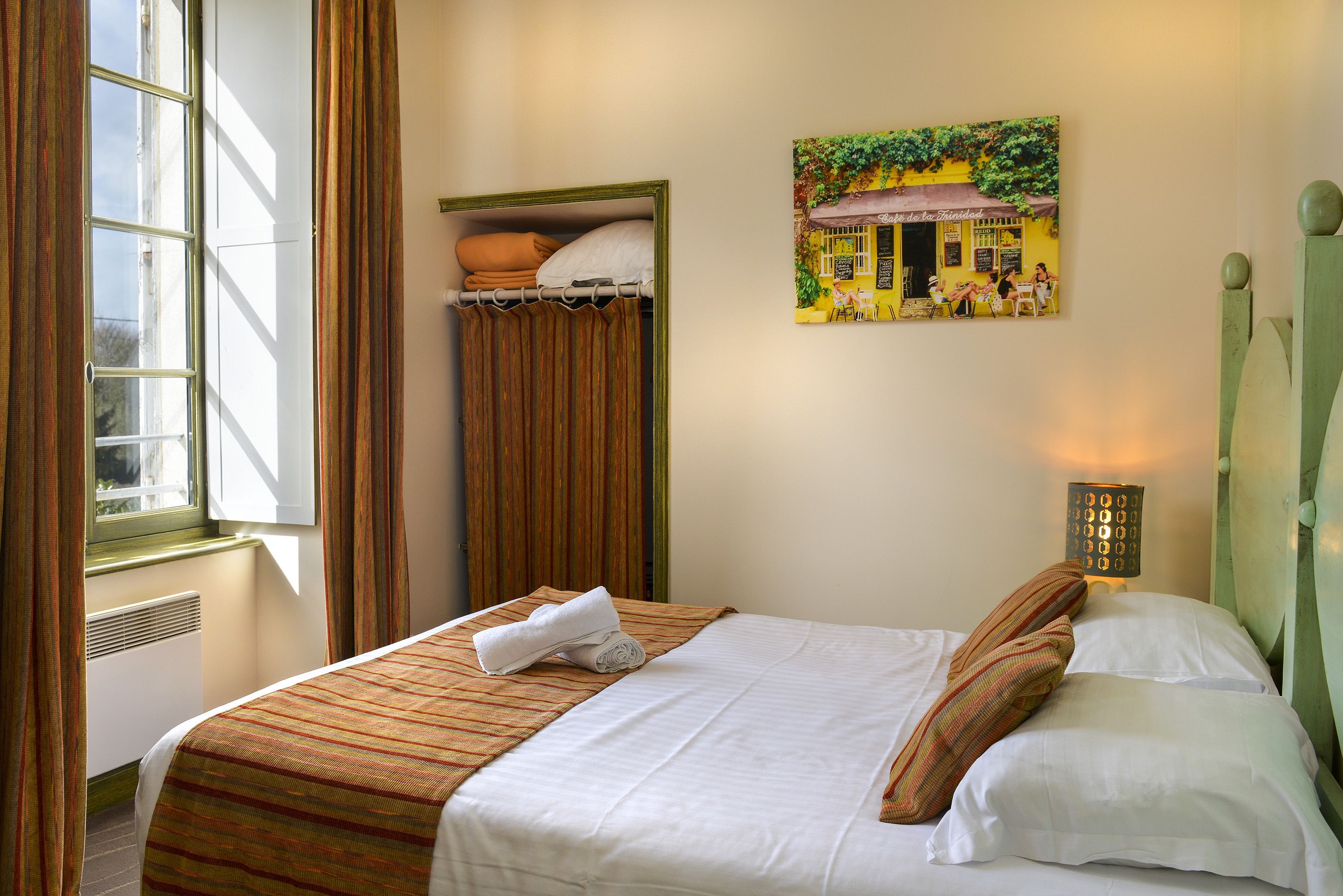 A neatly made bed with a striped bedspread, a pillow, and a rolled towel. There is a window with curtains, a radiator, and a wall-mounted light fixture.