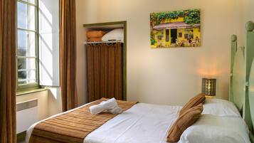 A neatly made bed with a striped bedspread, a pillow, and a rolled towel. There is a window with curtains, a radiator, and a wall-mounted light fixture.