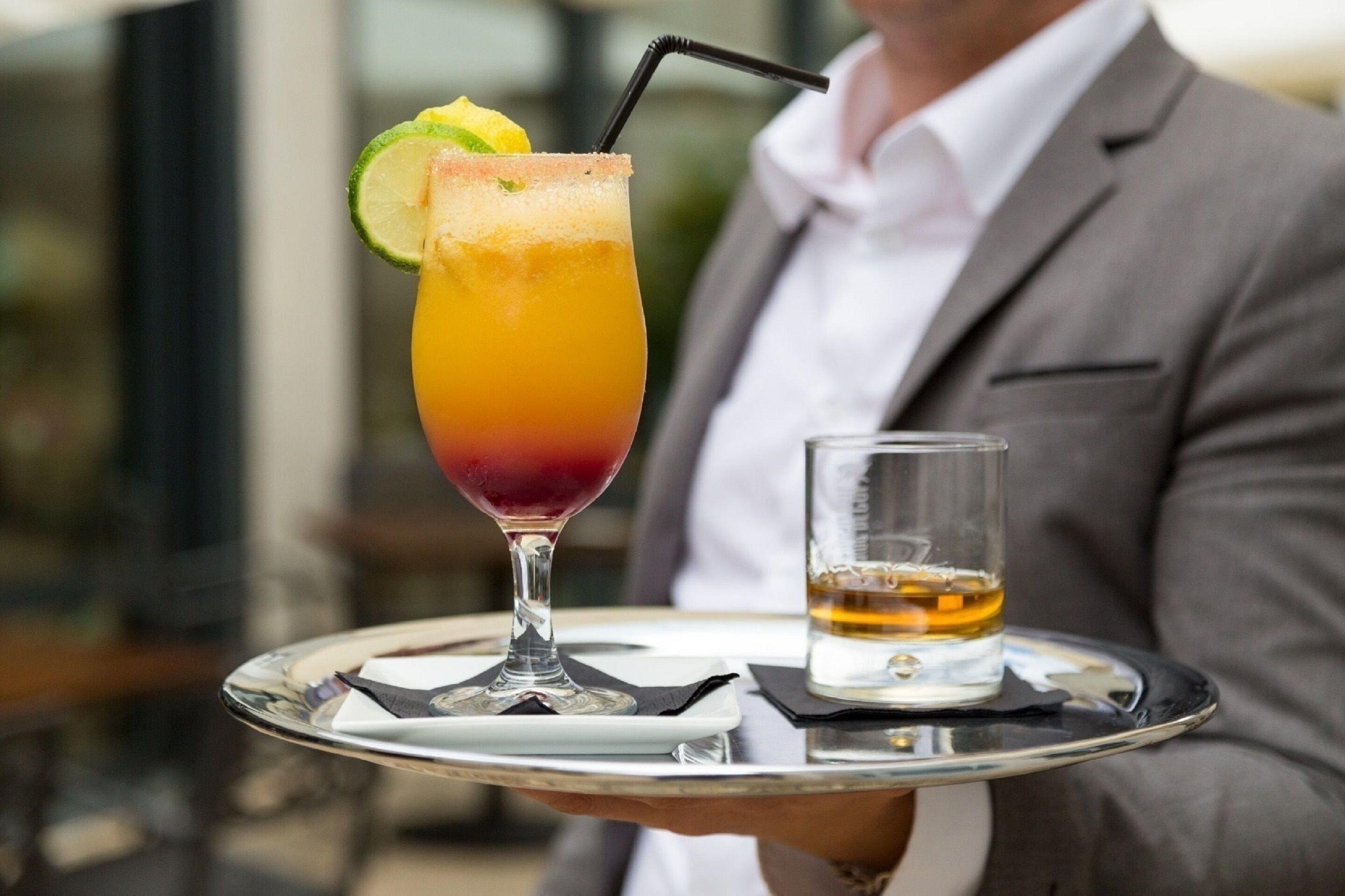 A cocktail in a tall glass with a lime garnish, served on a tray with a small glass of whiskey.