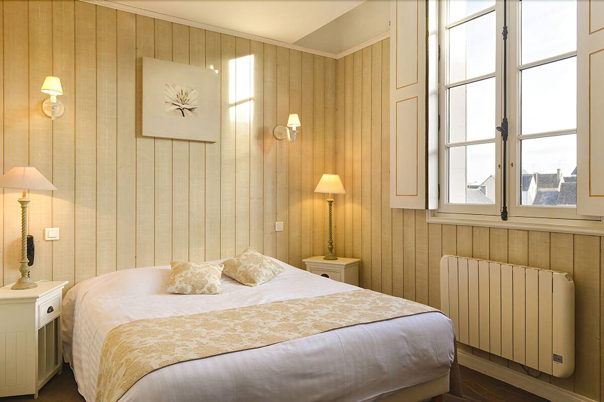 Een slaapkamer met een bed, twee bedlampjes, een aan de muur bevestigd schilderij, een raam met luiken en een radiator.