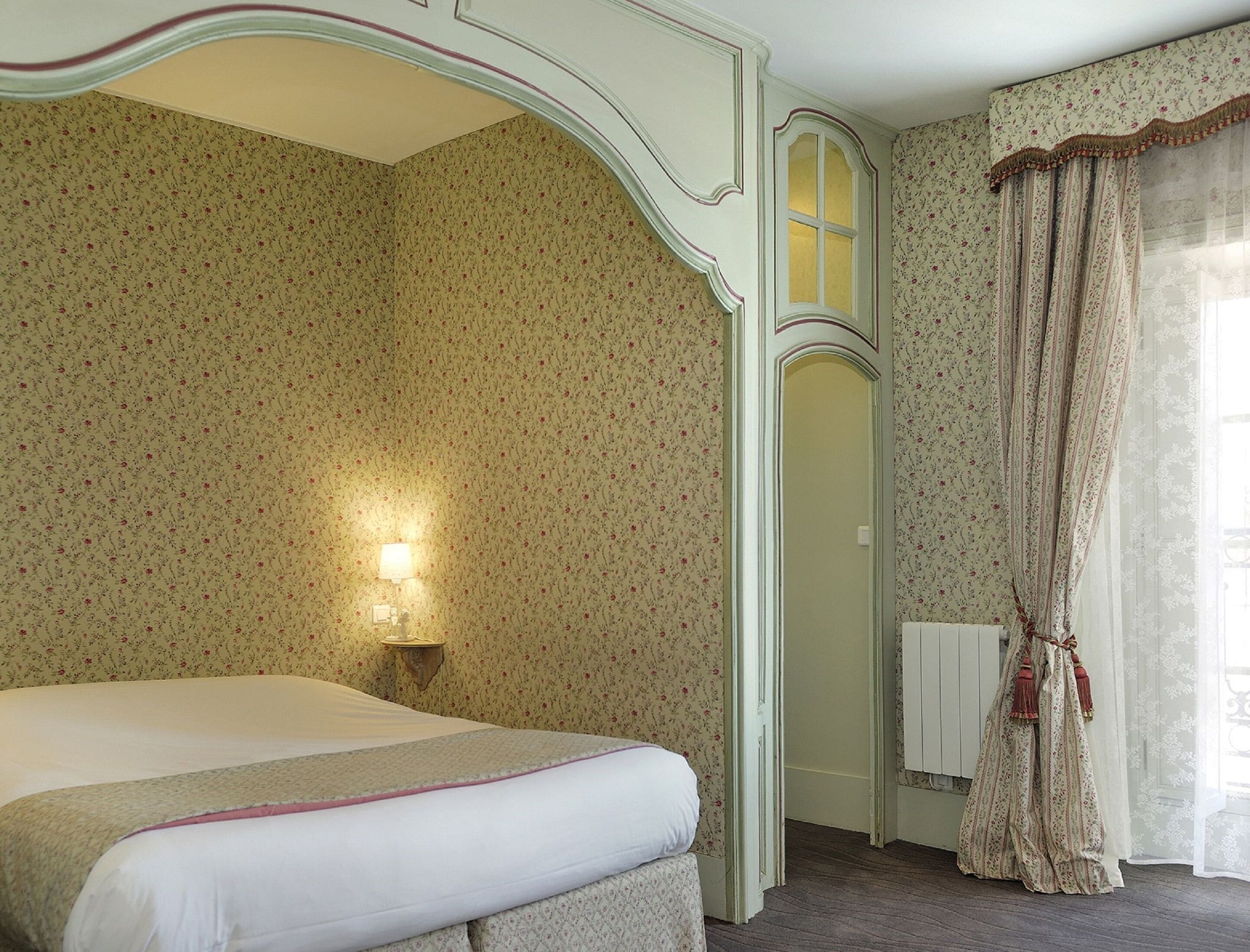 Un dormitorio con papel tapiz floral, una cama con colcha blanca, una mesita de noche con lámpara y una ventana con cortinas.