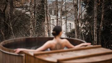 Una persona en un jacuzzi de madera rodeada de árboles, con un edificio al fondo.