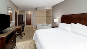 A hotel room with a bed, a desk with a flat-screen TV, a chair, a large mirror, a bathroom with a bathtub, and a nightstand with a lamp. - Hilton Garden Inn Augusta (Augusta)