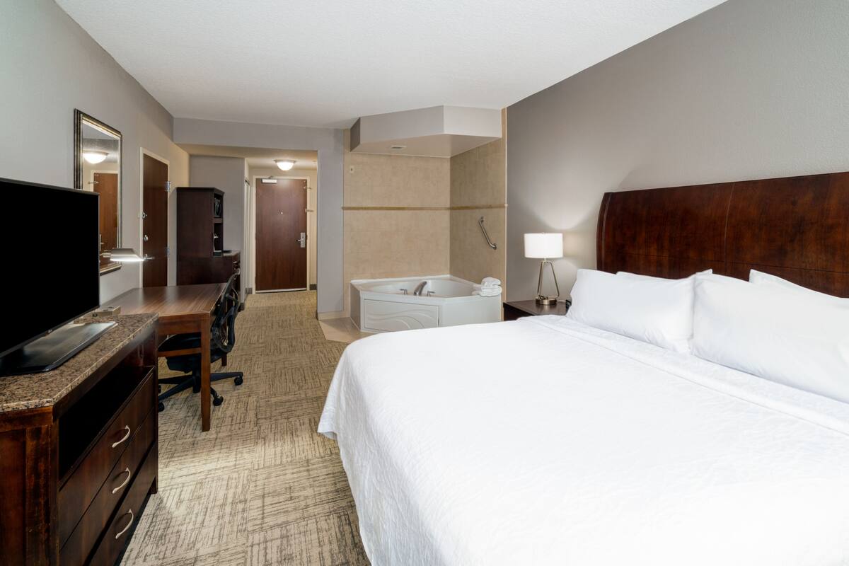 Quarto de hotel com cama, escrivaninha com televisão de tela plana, cadeira, espelho grande, banheiro com banheira e mesa de cabeceira com abajur.