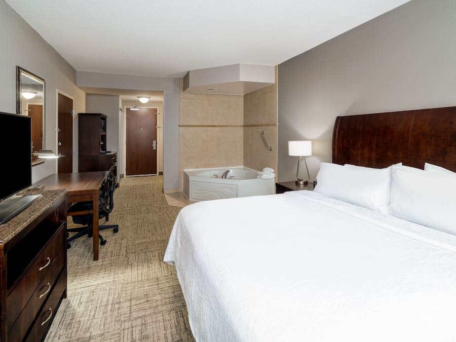 A hotel room with a bed, a desk with a flat-screen TV, a chair, a large mirror, a bathroom with a bathtub, and a nightstand with a lamp.