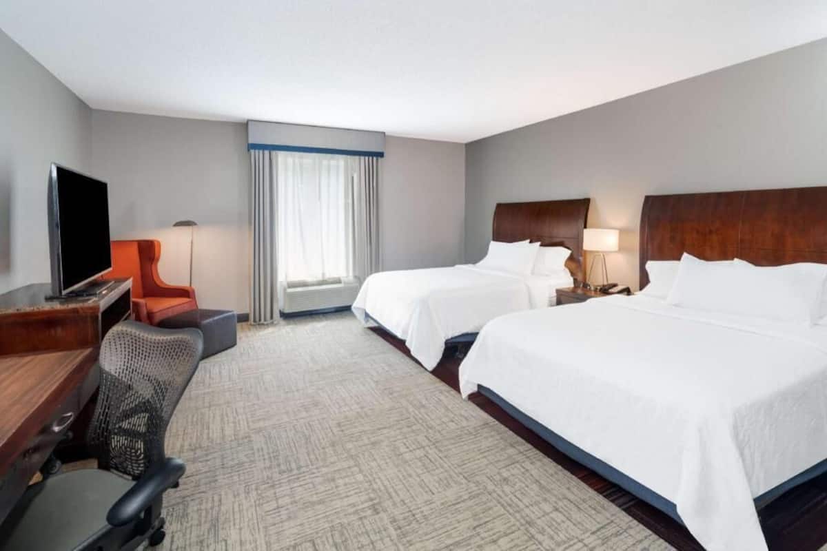 Quarto de hotel com duas camas, uma escrivaninha com televisão, uma cadeira e uma janela com cortinas.
