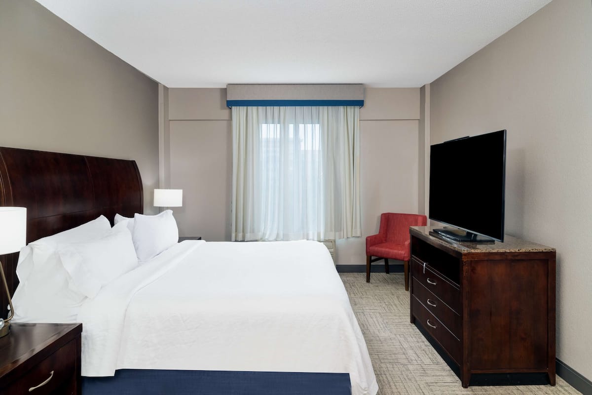 Quarto de hotel com uma cama grande, televisão em móvel de madeira, uma cadeira vermelha e uma janela com cortinas.