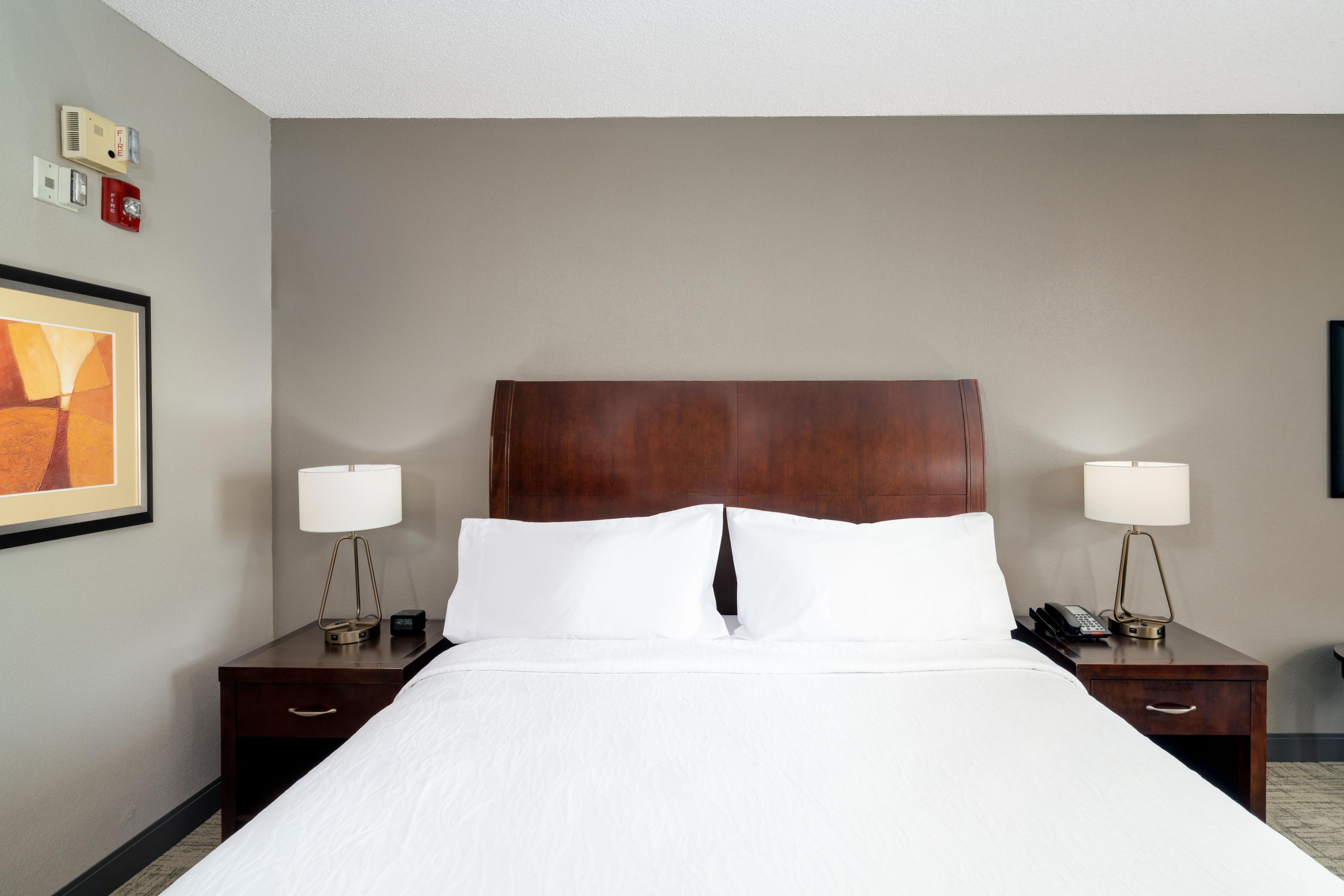 Une chambre d’hôtel avec un grand lit, deux tables de chevet avec des lampes, une horloge et un tableau encadré au mur.