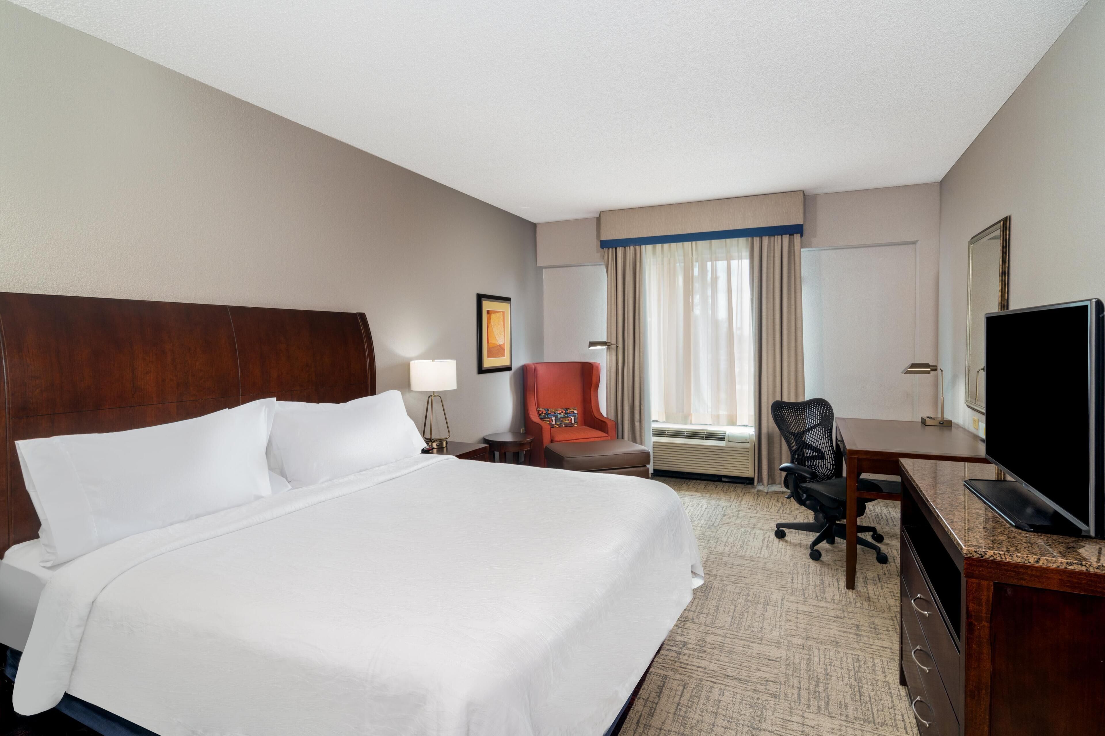Quarto de hotel com uma cama grande, uma escrivaninha com computador, uma cadeira, uma televisão e uma janela com cortinas.
