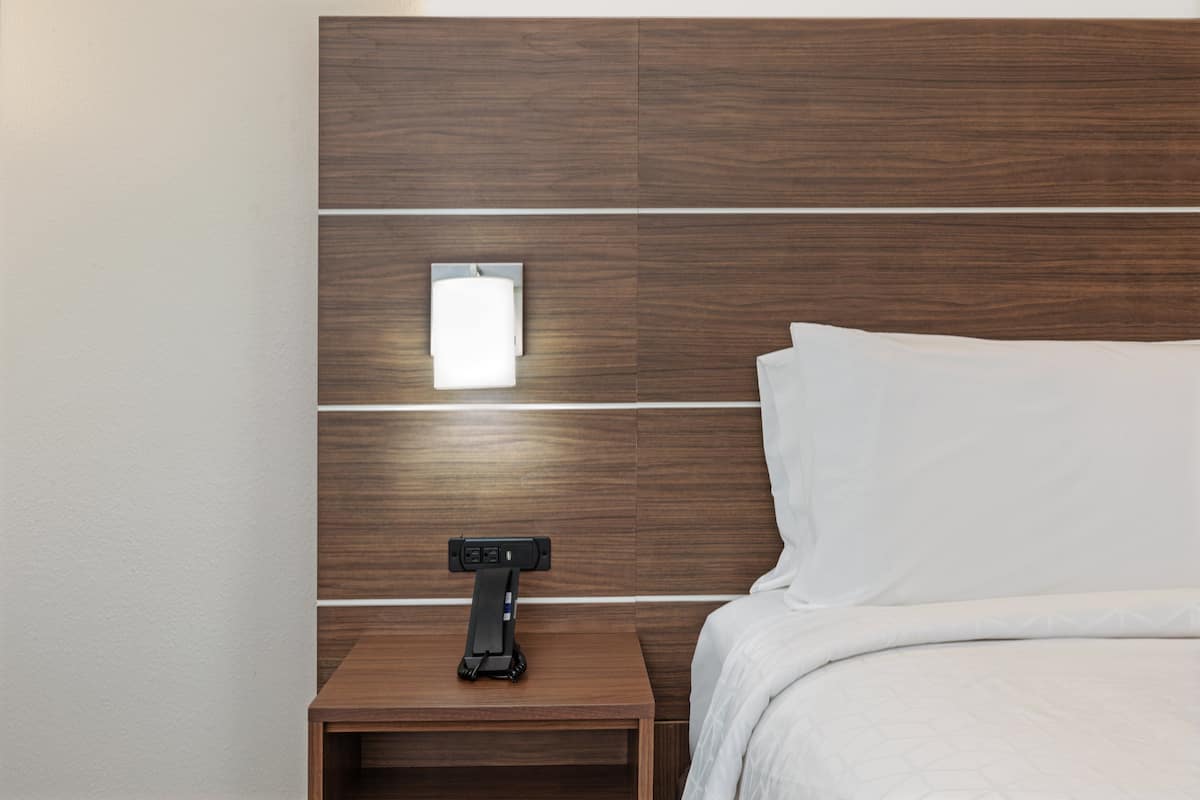A modern hotel room with a wooden headboard, a bedside table with a lamp, and a neatly made bed.