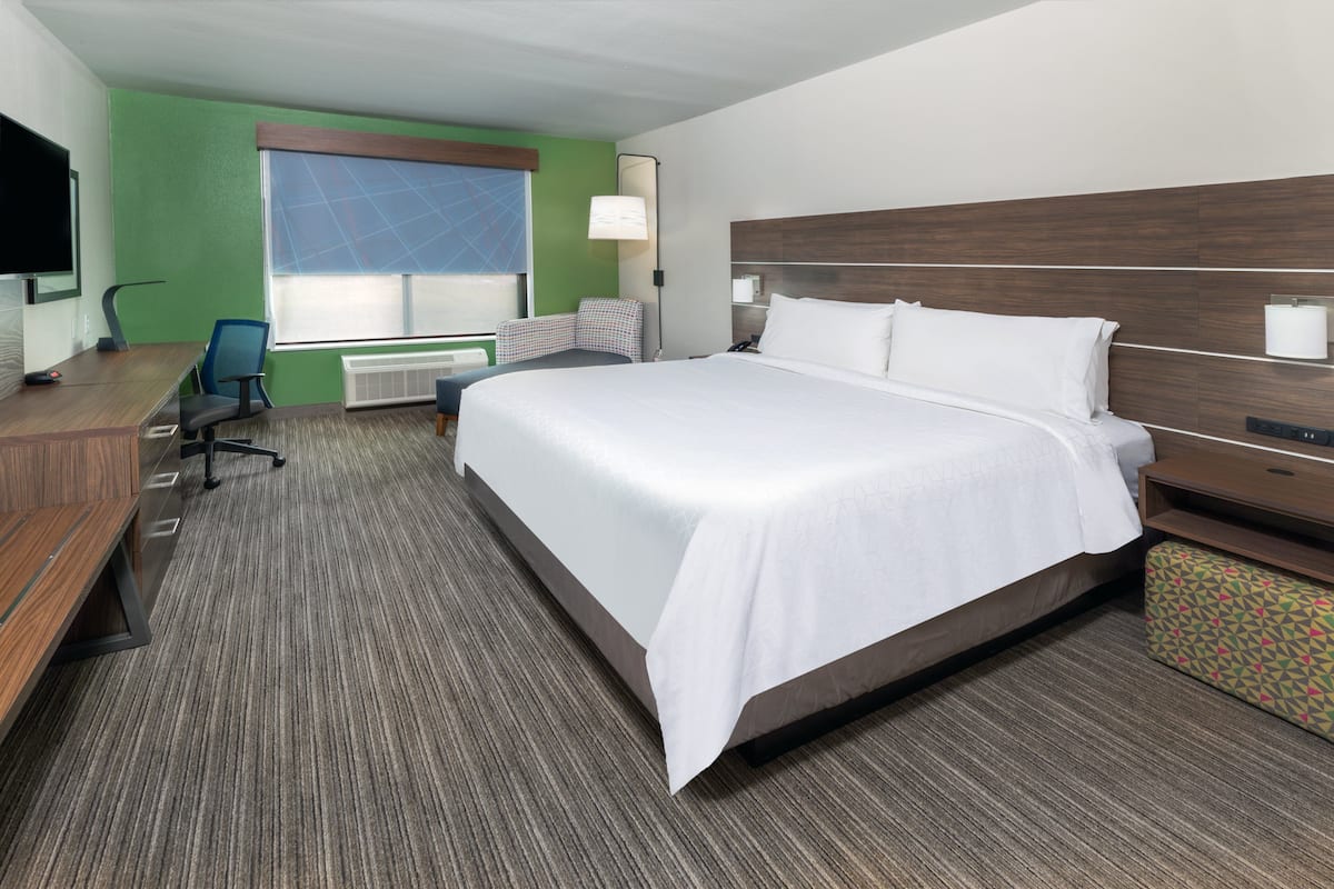 A hotel room with a large bed, a desk with a computer, a chair, a TV, and a window with blinds.