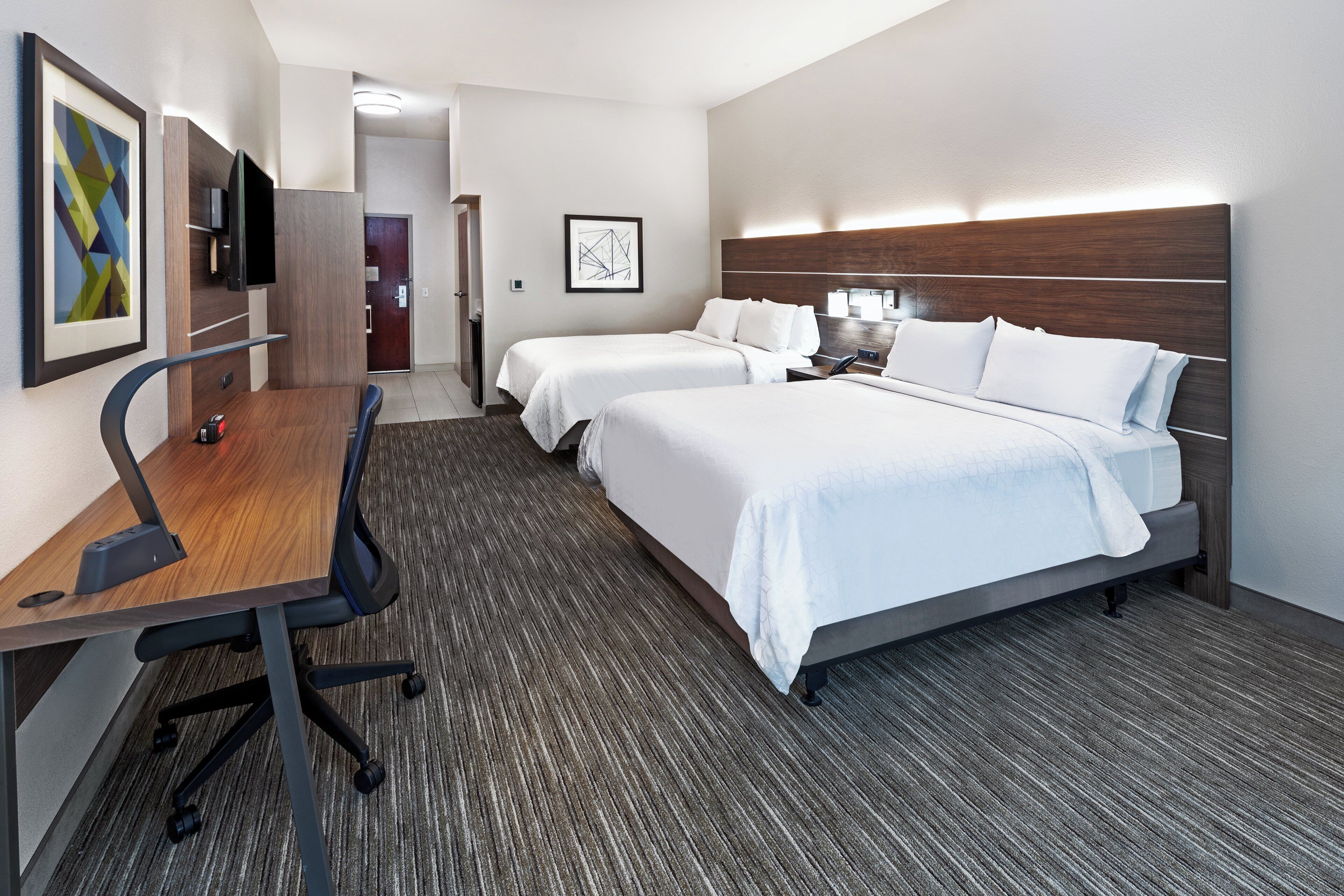 A hotel room with two beds, a desk, a chair, a TV, and artwork on the wall.