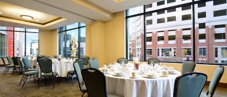 Un salón de conferencias con mesas redondas dispuestas para una reunión, amplias ventanas con vistas a un edificio y suelo cubierto de alfombra.