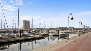 Ein Yachthafen mit vertäuten Booten, einer Uferpromenade und einem hohen Gebäude im Hintergrund.
