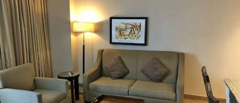 A hotel room with a sofa, armchair, coffee table, floor lamp, and a framed abstract painting on the wall.
