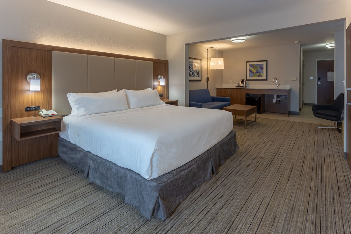 A hotel room with a large bed, bedside tables, a desk, a chair, and a kitchenette.