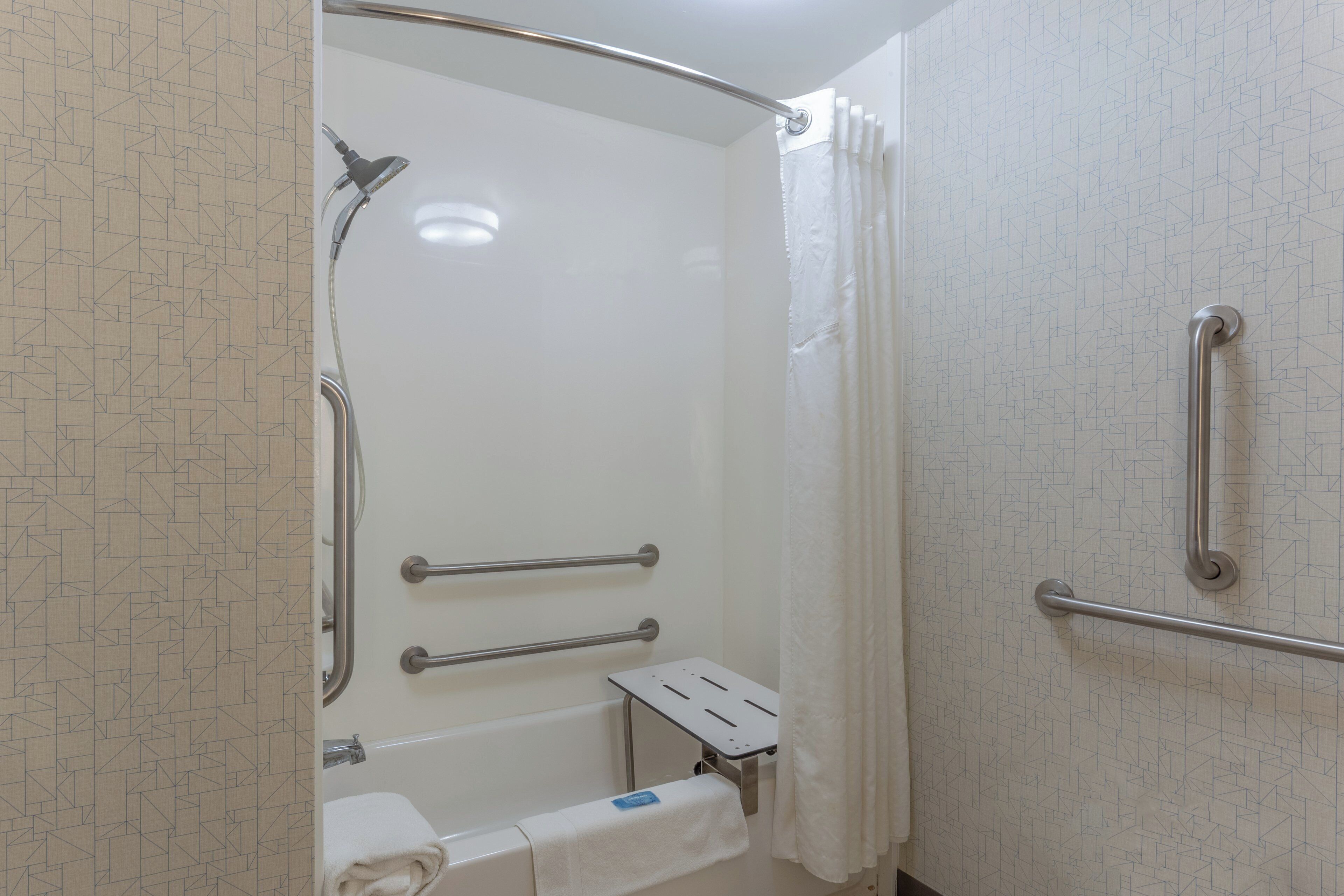 Baño con ducha a nivel, barras de apoyo, toallero y un pequeño banco.