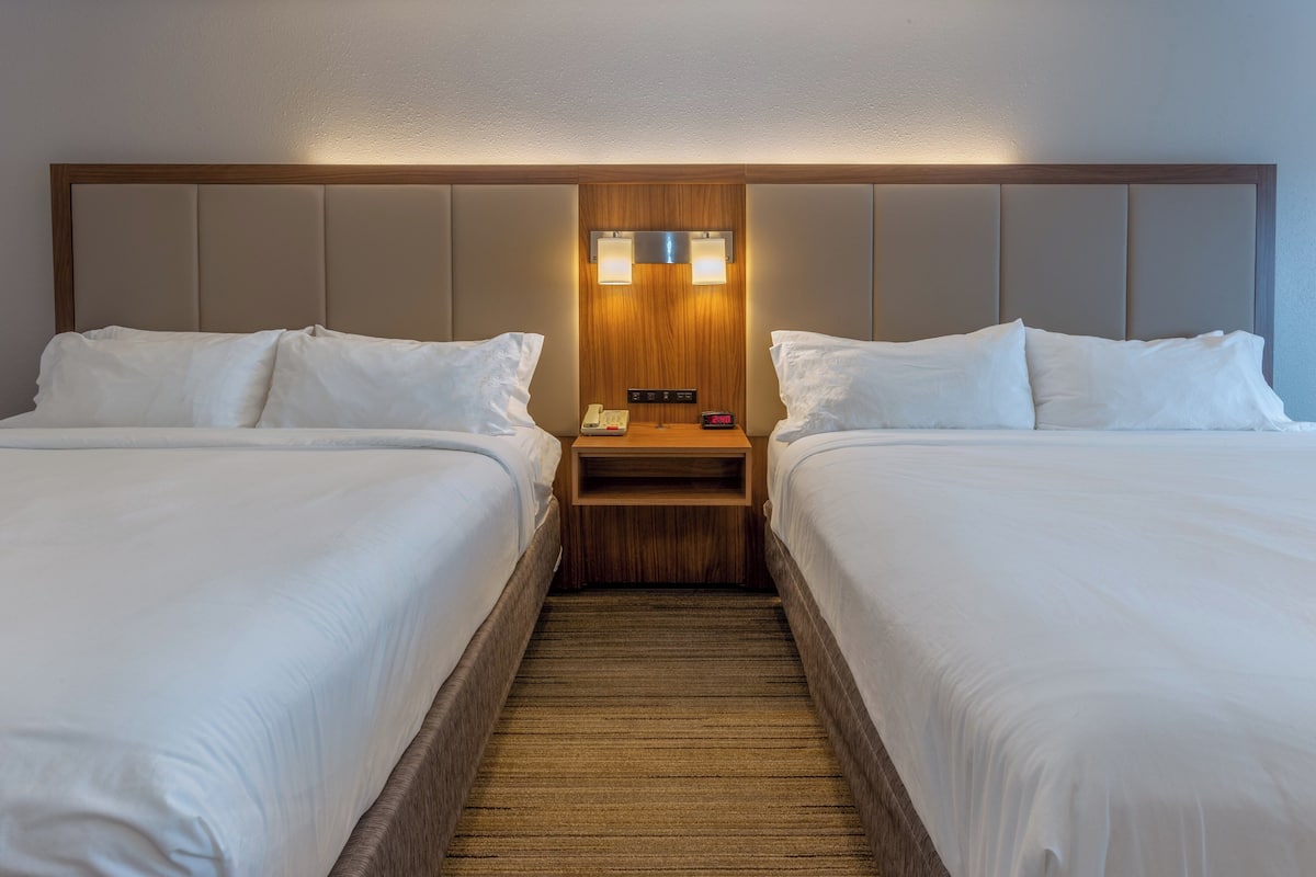 A hotel room with two beds, a wooden headboard, a nightstand with a phone and clock, and a carpeted floor.