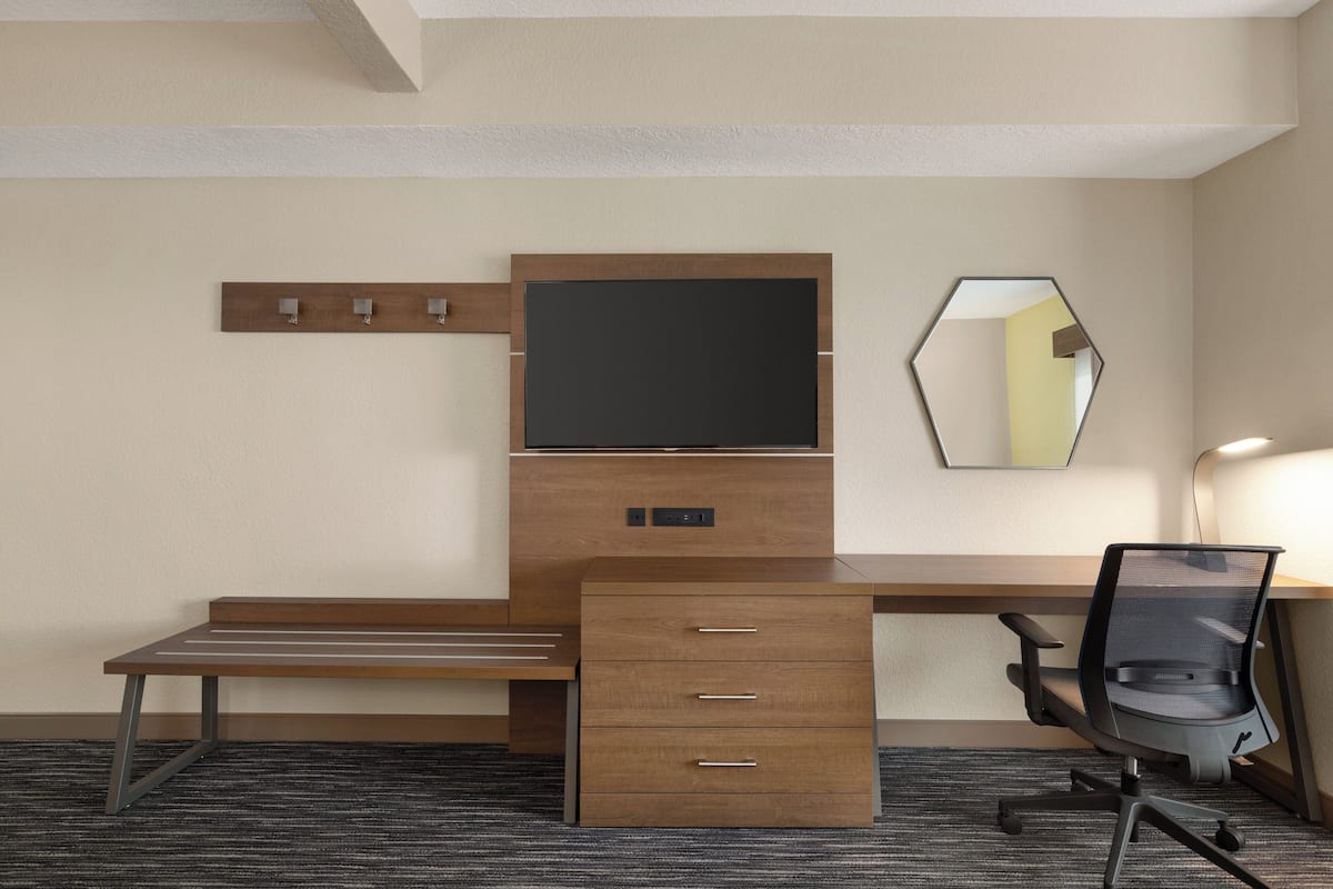 A hotel room with a wooden desk, a flat-screen TV, a mirror, and a chair.